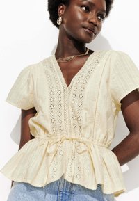 Blouse peplum en tissu crème clair avec des détails de broderie anglaise, des manches bouffantes courtes et une taille à cordon pour un ajustement réglable.