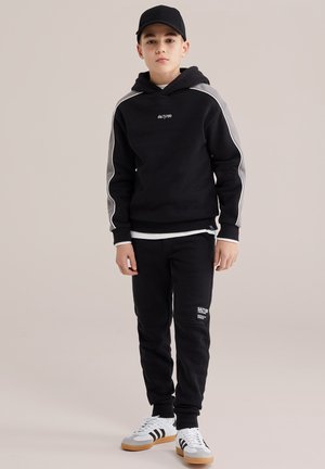 Junge trägt schwarzen Kapuzenpullover, schwarze Jogginghose mit Text-Patches, weiße Sneakers mit schwarzen Streifen und eine schwarze Baseballkappe vor schlichtem Hintergrund.