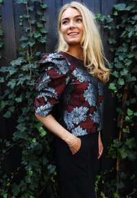 Majica s cvjetnim uzorkom u bordo i srebrnoj boji s napuhanim rukavima, uparena s crnim hlačama. Pozadina od zelenog bršljana dodatno ističe kontrast.