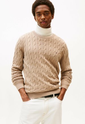 Beżowy sweter z warkoczowym splotem, z prążkowanymi mankietami i dołem, okrągłym dekoltem oraz małym logo. Model nosi białe spodnie z rękami w kieszeniach.