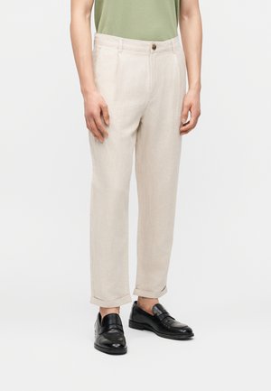 Mann iført beige bukser med oppbrett kombinert med svarte lærloafers, stående mot en ensfarget hvit bakgrunn.