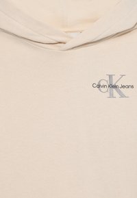Svetlo béžová mikina vyrobená z jemnej tkaniny, ktorá má vyšitý logo v sivej farbe, s nápisom "Calvin Klein Jeans" a dekoratívnym šnúrkami.