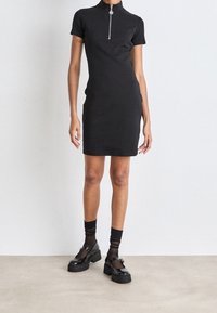 Czarna sukienka z krótkim rękawem, z wysokim kołnierzem i detalem zamka. Model nosi czarne masywne buty i przezroczyste czarne skarpetki, stojąc na jasnej podłodze.