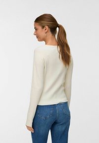Långärmad, krämfärgad tröja med texturerad tyg och rund halsringning, kombinerad med blåjeans. Synliga sömdetaljer på baksidan.
