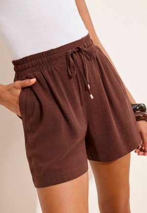 Femme portant un short marron à taille élastique avec un cordon, un haut blanc et des bracelets, une main dans la poche, sur un fond clair.