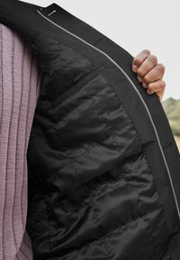 Veste isolante noire avec doublure matelassée, fermeture éclair argentée et poche intérieure, portée partiellement dézippée sur un haut côtelé violet.