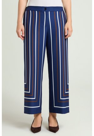 Donkerblauwe broek met wijde pijpen met verticale en vierkante witte en bruine strepen, gedragen met donkerbruine schoenen met spits toelopende neus en een witte top.