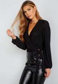 Sort indpakningsbluse med lange ærmer, rillet struktur, dyb V-hals og sidesnor detalje, parret med høj talje sorte faux læderbukser.