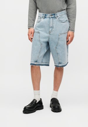 Φωτεινά γαλάζια τζιν σορτς μέχρι το γόνατο με μεγάλες μπροστινές τσέπες, φορεμένα με λευκές κάλτσες και μαύρα μοκασίνια.