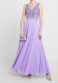 Lilat pitkä mekko, jossa paljetein koristeltu V-kaula-aukollinen yläosa, liehuva helma sekä hopeat korkokengät ja clutch-laukku. Sileä pinta kauttaaltaan.