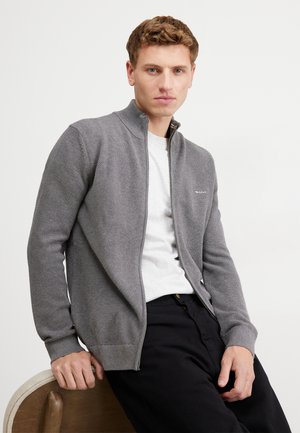 Cardigan grigio con zip, caratterizzato da un design a maglia testurizzata, polsini a costine e un logo sottile. Indossato sopra una t-shirt bianca e pantaloni neri.