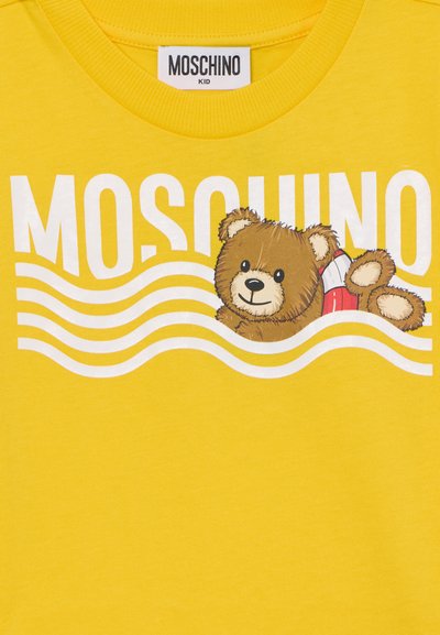 Žluté bavlněné tričko s ilustrací hnědého medvídka, červeným záchranným kruhem a bílým nápisem "MOSCHINO" s vlnovkovými vzory.