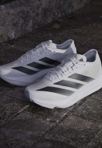 Ζευγάρι λευκά παπούτσια τρεξίματος Adidas με μαύρες ρίγες πάνω σε πέτρινη πλακοστρωμένη επιφάνεια.