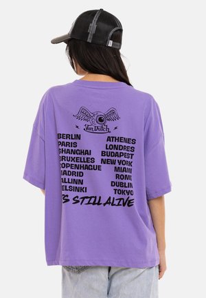 Personne portant un t-shirt violet oversize avec des noms de villes en noir et un graphique d'œil ailé dans le dos, associé à une casquette de baseball noire en mesh.