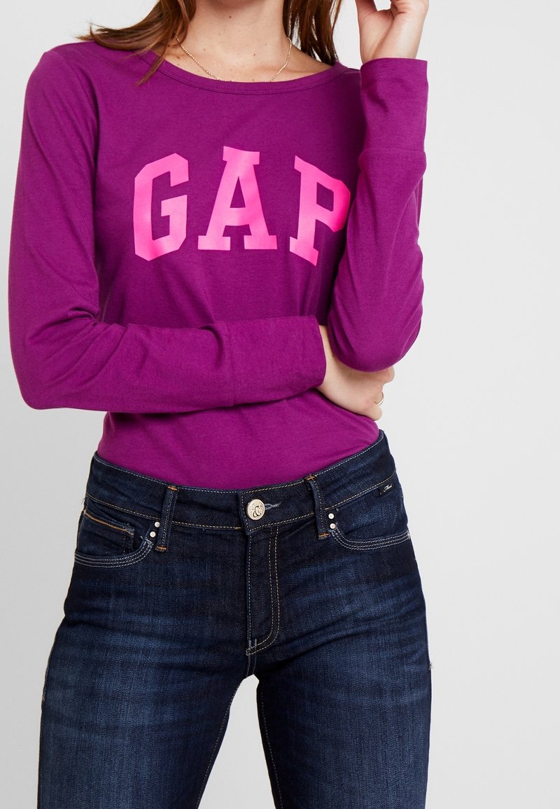 Långärmad lila skjorta med stora rosa "GAP" bokstäver, kombinerad med mörkblå jeans med detaljerad sömnad och knäppning.