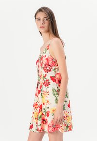 Abito floreale con uno sfondo di crema chiaro, decorato con fiori rossi e gialli vivaci. Presenta sottili spalline e una lunghezza fino al ginocchio.