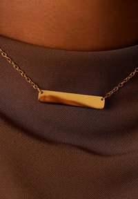 Gouden ketting met een gepolijste rechthoekige hanger, voorzien van een delicate ketting en geplaatst tegen een gestructureerde bruine stof.