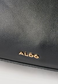Približek črnega teksturiranega usnjenega površja z majhno zlato ALDO blagovno znamko vtisnjeno blizu spodnjega roba.