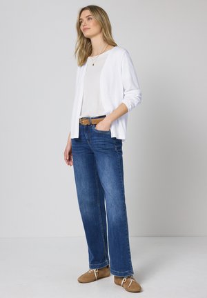 Kvinde stående iført hvid cardigan, hvid top, blå jeans, brun bælte og lysebrune snøresko mod en ensfarvet lys baggrund.