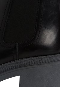 Zbliżenie czarnego skórzanego botka za kostkę z elastyczną boczną wstawką i teksturowaną krawędzią podeszwy.