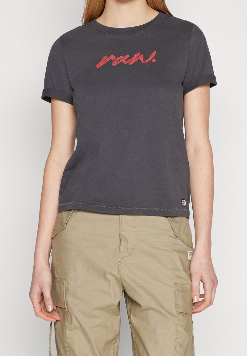 Femme portant un t-shirt gris foncé à manches courtes avec le texte rouge « raw. » et un pantalon cargo beige, debout devant un fond uni.