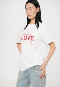 Biela tričko s krátkym rukávom s ružovou grafickou ilustráciou slova "LOVE". Tkanina má hladkú štruktúru a voľný strih.