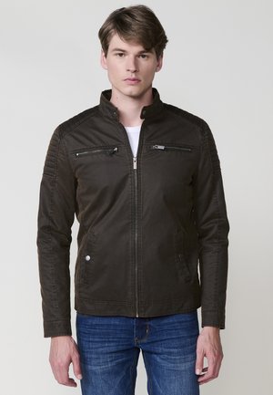 Chaqueta de color marrón oscuro con un alto cuello, acentos acolchados en los hombros, dos bolsillos con cremallera en el pecho y dos bolsillos laterales con botón a presión. Textura de tela suave.