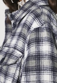 Primo piano di una persona che indossa un giubbotto a quadri grigio, bianco e nero con bottoni neri e una tasca a patta sulla spalla.