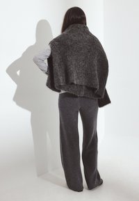 Nainen seisoo valkoisen nurkkaseinän edessä kasvoillaan, yllään tummanharmaat leveälahkeiset housut, vaaleanharmaa pitkähihaisen paita ja tumman pörröinen huivi hartioillaan.