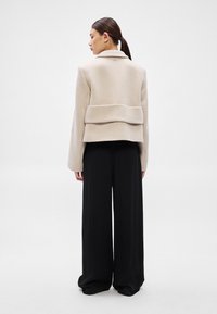 Μπεζ σακάκι από μείγμα μαλλιού με δομημένο σχέδιο, που διαθέτει φαρδιά γιακά και στρωμένη λεπτομέρεια στην πλάτη, συνδυασμένο με χαλαρό μαύρο παντελόνι.