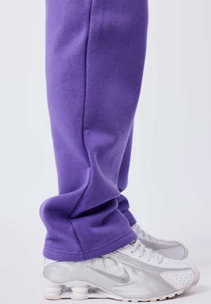 Persoon in paarse sweatpants en witte Nike sneakers met een unieke veerachtige zoolontwerp, staand op een effen achtergrond.