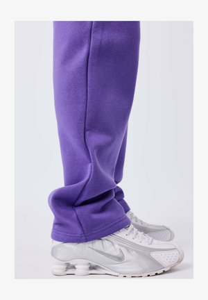 Persoon in paarse sweatpants en witte Nike sneakers met een unieke veerachtige zoolontwerp, staand op een effen achtergrond.
