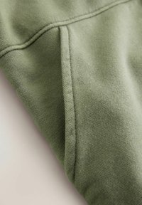 Tkanina oliwkowo-zielona o miękkiej fakturze, z ukośnym szwem i widocznymi detalami szwów. Materiał wydaje się przytulny i ciepły.