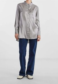 Camicia button-up in satin grigio chiaro con colletto, maniche lunghe e taschino sul petto, abbinata a jeans blu scuro e scarpe color crema.