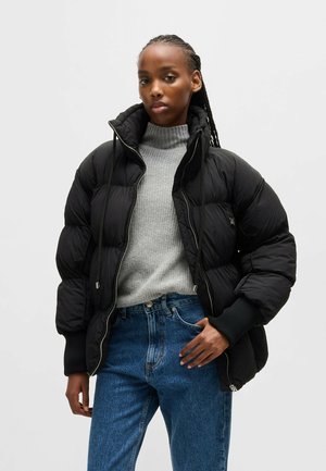 Schwarze Daunenjacke mit hohem Kragen, Reißverschluss und gerippten Bündchen. Getragen über einem grauen Rollkragenpullover und blauen High-Waist-Jeans.