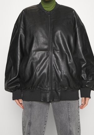 Osoba nosząca oversize czarną skórzaną kurtkę typu bomber na zielonym golfie i szarych dżinsach, stojąca na jednolitym tle.