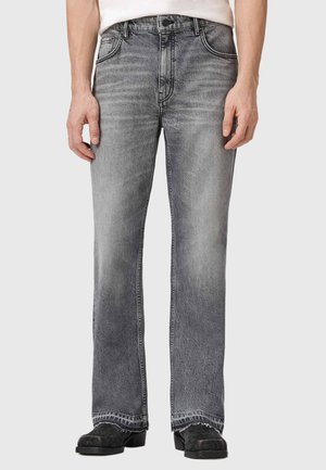 Man draagt grijze vervaagde rechtbenige spijkerbroek en zwarte schoenen, staat met ontspannen handen langs zijn lichaam tegen een effen achtergrond.