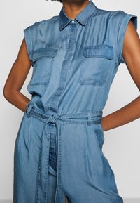 Tuta in denim di blu chiaro con maniche corte, due tasche anteriori, colletto e cintura in tessuto legata in vita.
