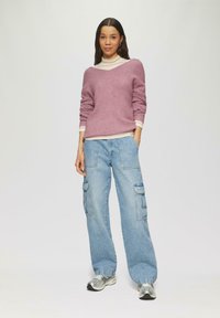 Lichtpaarse gebreide trui met een diepe V-hals, gelaagd over een crèmekleurige turtle neck. gecombineerd met loszittende lichtblauwe cargojeggings en zilveren sneakers.