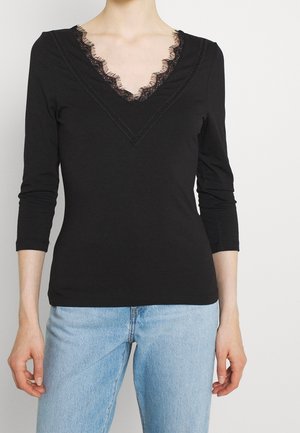 Chemise noire à manches longues avec un décolleté en V profond et des garnitures en dentelle. Fabriquée en tissu doux, design ajusté, et accompagnée de jeans bleu clair.