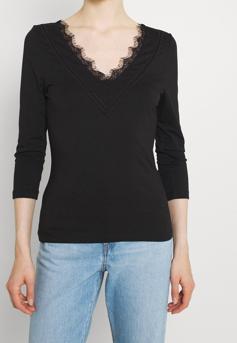 Maglietta nera a maniche lunghe con scollatura a V profonda e bordo in pizzo. Realizzata in tessuto morbido, con un design aderente, abbinata a jeans azzurri chiari.