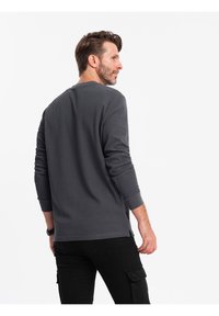 Maglietta grigia a maniche lunghe con scollatura rotonda, spacchi laterali, polsini a costine e una finitura testurizzata, indossata con pantaloni cargo neri.