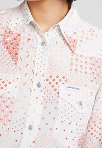 Camisa blanca de botones con patrones geométricos y florales rojos, botones a presión plateados, cuello puntiagudo y bolsillo en el pecho etiquetado "Calvin Klein Jeans".