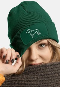 Dark green knitted beanie with a textile patch featuring a white outline of a dog. Fabric is ribbed, offering a textured appearance.