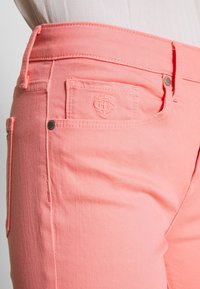 Svetlé korálové džíny s prošívanými kapsami a stříbrnými kovovými prvky. Tkanina má hladkou denimovou strukturu a přiléhavý střih.
