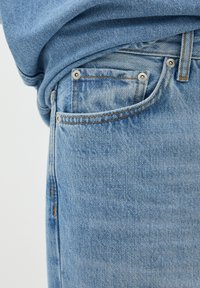 Los jeans de mezclilla azul claro presentan un leve desgastado, diseño de cinco bolsillos con remaches y costuras naranjas contrastantes a lo largo de las costuras.