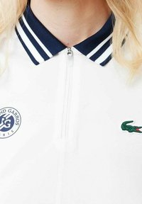 Polo shirt blanc avec un col zippé, un col rayé bleu marine et blanc, et des logos brodés : un rond et un crocodile. Texture piqué.