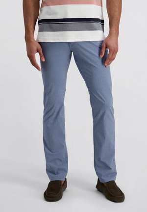 Man die lichtblauwe slim-fit broek, bruine loafers en een gestreept shirt met korte mouwen draagt, staand tegen een effen achtergrond.