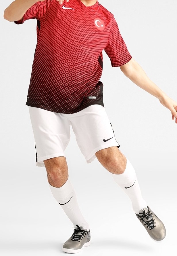 Maillot rouge et noir avec un motif en grille et un emblème de la Turquie, associé à un short blanc. Des baskets grises et des chaussettes blanches complètent la tenue.