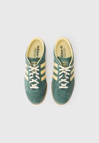 Zielone zamszowe sneakersy z trzema żółtymi paskami, białymi sznurowadłami i gumową podeszwą. Złote logo na języku.